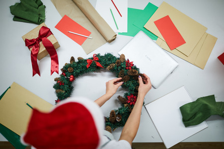 Directly above view of unrecognizable craftsperson in Santa hat standing at desk with papers and laptop and decorating Christmas wreath with pine conesの写真素材