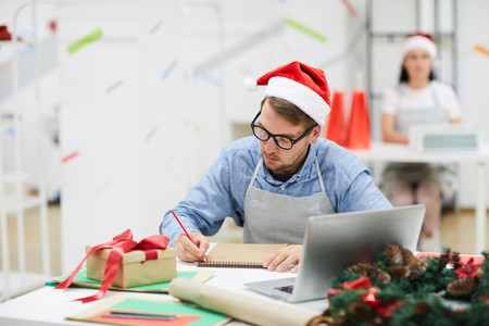 Serious concentrated young male designer in Santa hat sitting at table with laptop and gift box and planning work in creative studioの写真素材