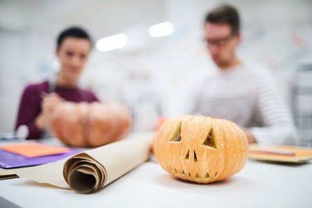 Close-up of carved Halloween pumpkin and parchment paper roll on table, crafts people working in backgroundの写真素材