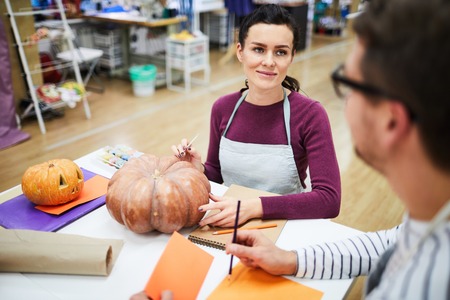 Content young colleagues of crafting studio sitting at table and chatting while working on design of Halloween pumpkinsの写真素材