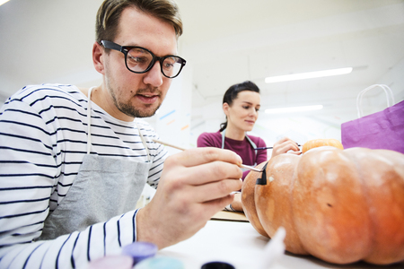 Content concentrated young man in glasses sitting at table and enjoying painting food in art studio, he drawing black eyes on pumpkinの写真素材