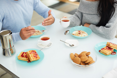 Tasty breakfast with lover: close-up of unrecognizable couple sitting at dining table and eating porridge and drinking tea while discussing interesting topic, Belgium waffles and cookies on tableの写真素材