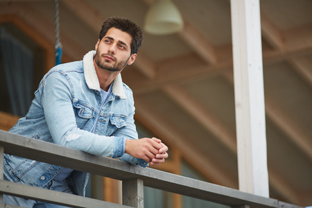 Content dreamy young man in warm denim jacket leaning on railing and looking away while resting on balcony of countryside houseの写真素材