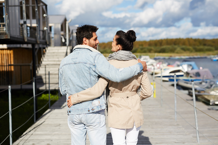 Rear view of young couple in casual clothing embracing each other and enjoying stroll on warm autumn day while walking on pierの写真素材
