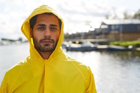 Portrait of serious handsome young bearded man in hood wearing waterproof coat standing at dockの写真素材