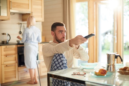 Young bearded man sitting at kitchen table and switching channels on TV with remote control while his wife making breakfastの写真素材