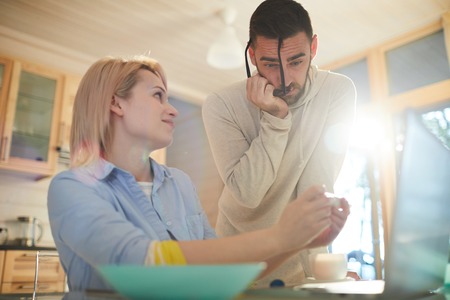 Young Caucasian woman sitting at kitchen table and showing pregnancy test with positive result to her husband, man looking surprised and embarrassedの写真素材
