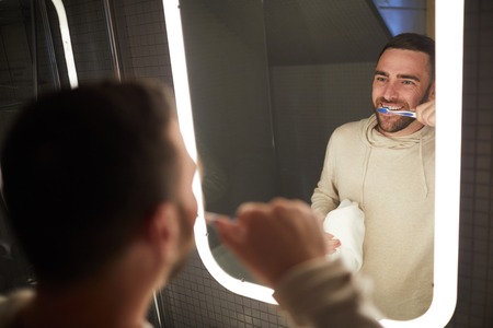 Reflection of young bearded Caucasian man brushing his teeth with toothbrush and looking at himself in bathroom mirrorの写真素材