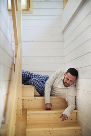 Young Caucasian man in homewear lying on wooden staircase at his house and wincing at pain after falling from high distanceの写真素材