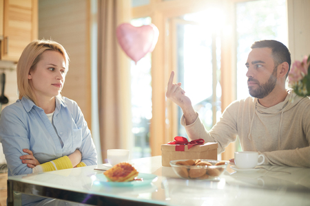 Displeased young Caucasian woman sitting at kitchen table with gift box while her frustrated husband giving her middle fingerの写真素材
