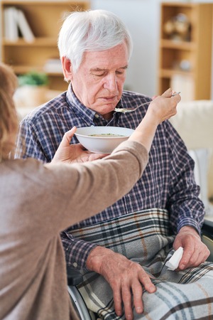 Aged female holding spoon and plate with soup by her disable patient or husband mouth while feeding...
