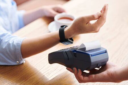 Close-up of unrecognizable businesswoman using smartwatch for wireless payment in modern cafe,...