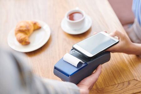 Close-up of restaurant guest sitting at table and using smartphone while paying through mobile...