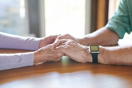 Affectionate and devoted senior spouses holding by hands while sitting in front of each other by...