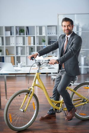 Young successful businessman sitting on bicycle in officeの写真素材