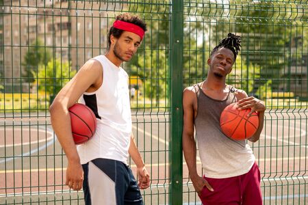 Two active guys in sportswear standing by fence surrounding basketball court