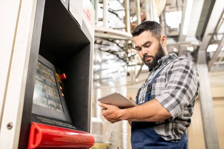 Bearded young worker of polymer production looking through data in touchpad