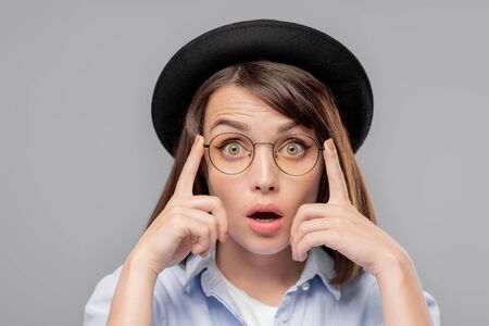 Young astonished female in hat and eyeglasses looking at you