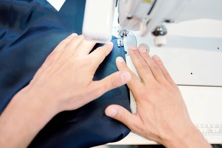 Human hands holding dark blue fabric while sewing clothing item of new fashion collection by machine