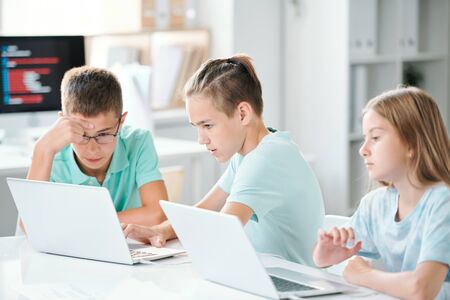Cute schoolchildren sitting by desk in classroom while browsing in the netの写真素材