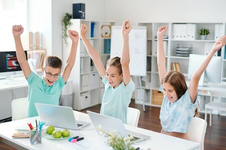 Cheerful middle school students with raised hands looking at laptop displaysの写真素材