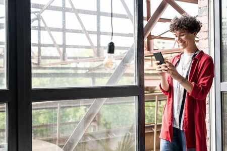 Young cheerful woman in casualwear standing by open window and using smartphone