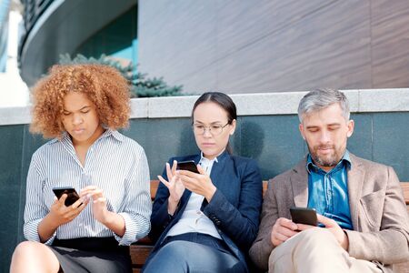 Row of mobile employees sitting along wall and scrolling in their smartphones