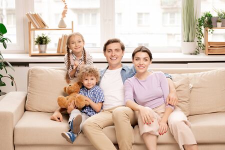 Four family members in casualwear sitting on couch by window in living-room