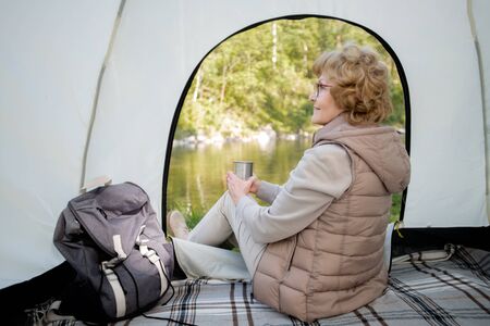 Active mature female backpacker with hot drink sitting in tentの写真素材