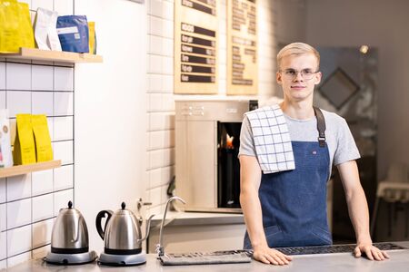 Young male barista in uniform standing by table with working equipmentの写真素材
