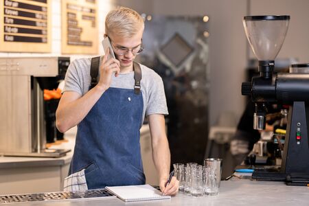 Young serious waiter or barista writing down order of client on the phoneの写真素材