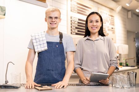 Young cheerful and friendly workers of cafe standing by counter