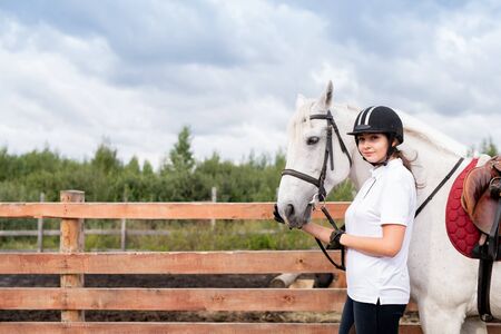 Young woman in equestrian outfit and white racehorse moving along wooden fenceの写真素材