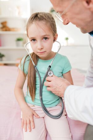 Puzzled little girl listening to her heartbeat in stethoscope during examinationの写真素材