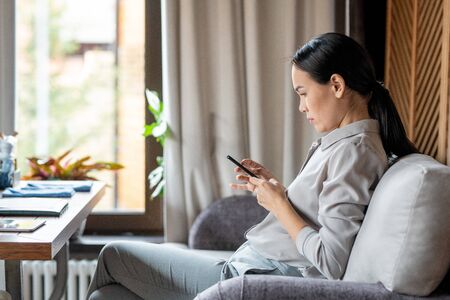 Young casual woman with smartphone sitting on couch in restaurant and messagingの写真素材