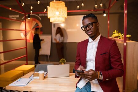 Young African businessman with mobile phone standing by table in front of cameraの写真素材
