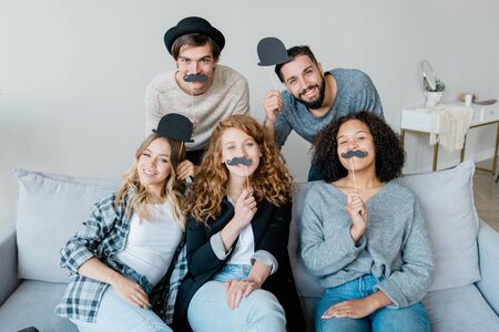 Joyful guys and girls holding moustaches and hats on sticks by mouths and headsの写真素材