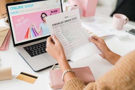 Young female customer holding and reading checklist paper over laptop keypad