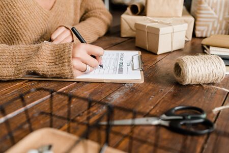 Young woman putting ticks by ordered goods in checklist while sitting by wooden table among packed boxesの写真素材