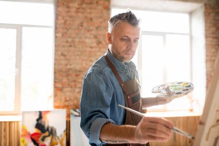 Inspired man with palette and paintbrush standing in front of easel and painting