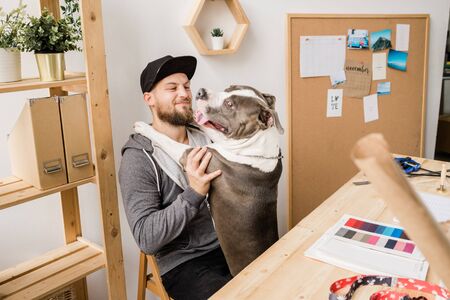 Happy young casual man sitting on chair by wooden table and playing with his petの写真素材