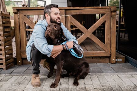 Happy young man sitting on squats and embracing his cute brown purebred dogの写真素材
