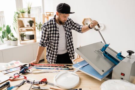 Young creative man printing or sticking decor on one of pet collar workpieces while using technical equipmentの写真素材