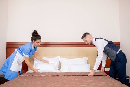Young porter and chamber maid bending over bed while making it in the morningの写真素材