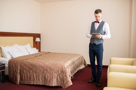 Young elegant porter using digital tablet while standing by bed in hotel roomの写真素材