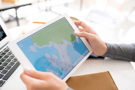 Hands of young female student holding digital tablet with map on display while preparing homework or...