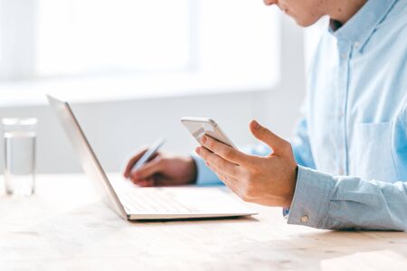 Hand of young man holding smartphone over table while sitting in front of laptop and making working...