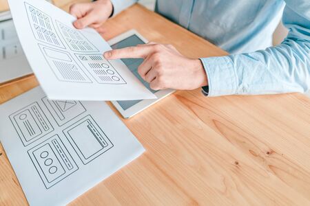 Hand of young interface designer pointing at program interface on one of papers while working over...