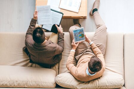 Overview of two young male economists sitting on couch, analyzing diagram and looking through financial papersの写真素材