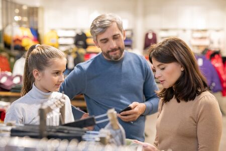 Girl and her parents standing by rack with casualwear in clothing departmentの写真素材